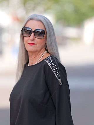 Doris Streich Pulli 279184 black: Frau mit langen grauen Haaren trägt ein schwarzes Shirt mit Glitzereinsatz, Zipper an der linken Schulter, kombiniert mit Sonnenbrille und Perlenkette.