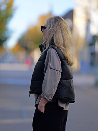 Frau trägt die Wendeweste OFF#DLY Anita black mit Stehkragen und seitlichen Taschen. Die Weste wird über einem schwarzen Oberteil getragen. Accessoires wie Sonnenbrille und Handtasche ergänzen den Look.