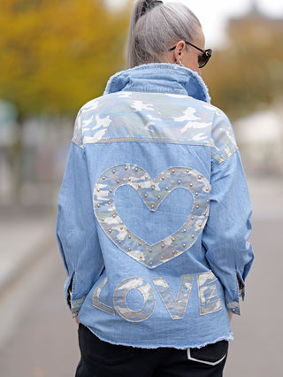 Frau trägt das OFF#DLY Denimshirt Camo mit Herz-Patch auf dem Rücken. Das Hemd hat Nieten und aufgesetzte Taschen, passend für Street-Fashion und Casual-Looks.