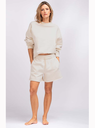 Frau trägt True Religion Super Crew Sweat palbeige, einen lässigen, kurz geschnittenen Sweater mit überschnittener Schulter und Washed out Look, kombiniert mit weißen Shorts.