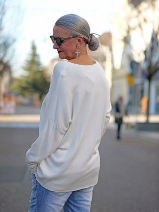 Frau in weißem OFF#DLY Pulli Yara creme mit V-Ausschnitt, oversized Passform. Sie trägt Sonnenbrillen und Kreuzohrringe, in lässigem Straßenstil mit Jeans.