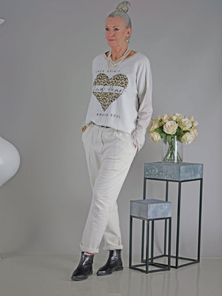 Frau trägt das OFF#DLY Tee Free Spirit beige mit Herz-Leopardenmuster, lange Ärmel und Rundhals, vor einer Vase mit weißen Rosen. Kombiniert mit weißen Hosen und schwarzen Stiefeln.