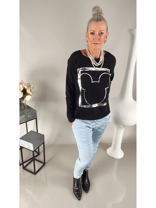 Frau trägt OFF#DLY Longsleeve Frame schwarz mit Silberprint und Rundhalsausschnitt, kombiniert mit blauen Hosen. Das Oberteil hat eine gerade Passform und ist aus Baumwoll-Elastan-Mix.