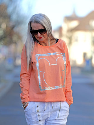 OFF#DLY Longsleeve Frame apricot: Frau mit Sonnenbrille trägt ein oranges Longsleeve mit Rundhalsausschnitt und Silberprint, gerade Passform. Ideal für lässige Street-Fashion.