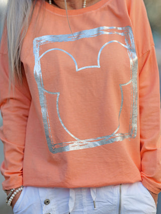 Eine Person trägt das OFF#DLY Longsleeve Frame apricot mit einem Cartoon-Mauskopf-Design auf dem Shirt, das einen Rundhalsausschnitt und lange Ärmel hat.