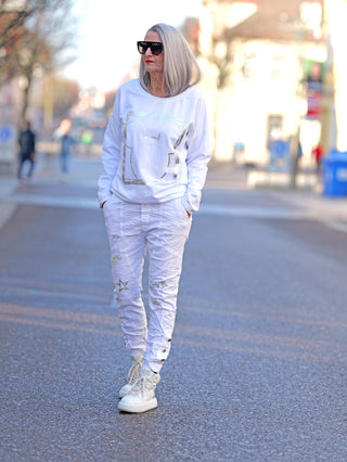 Frau trägt OFF#DLY Longsleeve Frame weiß mit Silberprint und Rundhalsausschnitt auf der Straße, kombiniert mit weißen Hosen und Sneakern, graues Haar und Sonnenbrille.