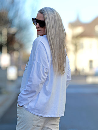 Frau mit langen blonden Haaren trägt OFF#DLY Tee Free Spirit weiß, ein Longsleeve mit Rundhalsausschnitt und Herz-Print, während sie Sonnenbrillen trägt.