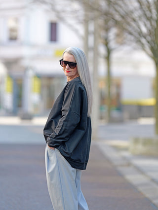 Eine Frau mit langen grauen Haaren trägt die OFF#DLY Sweat Schuh black, ein schwarzes Rundhals-Sweatshirt mit Glanzprint, leicht oversized, während sie in die Kamera schaut.