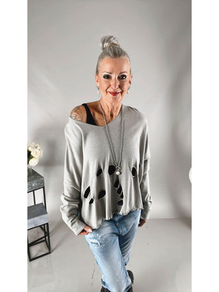 Philomena Pulli 115 silver: Frau trägt einen silbernen, grauen, boxy geschnittenen Pulli mit Löchern, hinten länger geschnitten. Kombiniert mit blauer Jeans und einer Halskette.