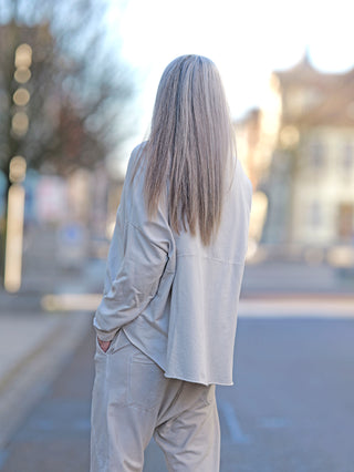 Das Bild zeigt das OFF#DLY Longsleeve Rita stone, getragen von einer Frau mit langen Haaren auf der Straße. Es hat einen Rundhals und einen vorne angebrachten Patch.