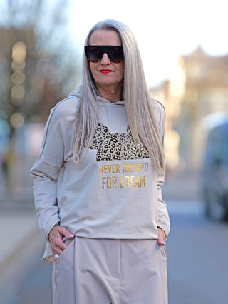 Frau mit langen Haaren trägt eine Sonnenbrille und ein OFF#DLY Hoody Tee Dream beige mit Goldprint, gerade Passform, geeignet für Größen 34 bis 42, fotografiert im Street-Fashion-Stil.