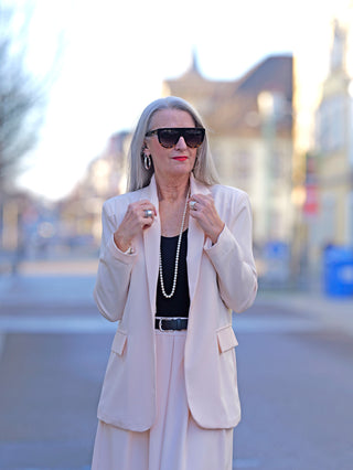 Eine Frau trägt den OFF#DLY Blazer Bibi apricot, einen leichten, gefütterten Ein-Knopf-Blazer mit angedeuteten Seitentaschen, kombiniert mit Sonnenbrille und einem Perlenhalsband.