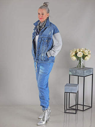 Frau trägt die OFF#DLY Denim Jacket Conny midblue/grey mit Sweatärmeln, steht vor einem Blumenstrauß in einer Vase. Die Jacke hat eine oversized Passform.