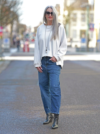 Frau auf der Straße trägt True Religion Hope Boe Leg Jeans, Wide Fit, mit Destroy-Waschung. Kombiniert mit weißem Pullover und Stiefeln, repräsentiert lässige Straßenmode. Ideal für moderne, bequeme Looks.