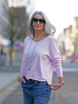 Eine Frau trägt den True Religion V-Neck Boxy pink Pullover, kombiniert mit blauen Jeans und Sonnenbrille, präsentiert den verkürzten, boxy Schnitt des Pullovers.