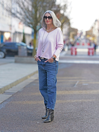 Frau trägt True Religion V-Neck Boxy pink Pullover auf Gehweg, betont lässigen, verkürzten Schnitt. Pullover aus Baumwolle, kombiniert mit blauer Jeans, vermittelt modernes Straßenmodegefühl.